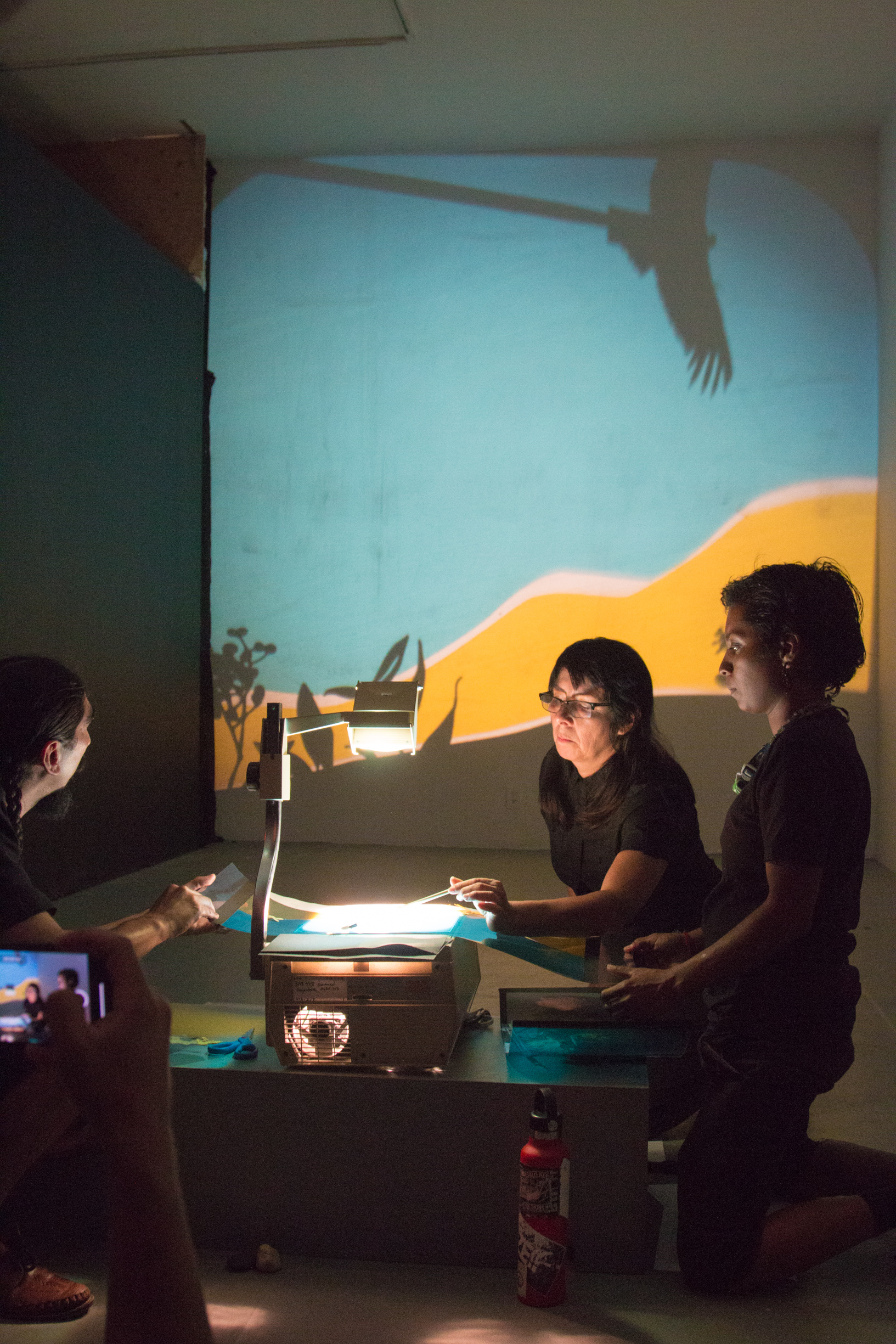 three people gather around an old-school projector. A colorful collage with yellow, blues and shadow puppets is projected onto the wall behind them.